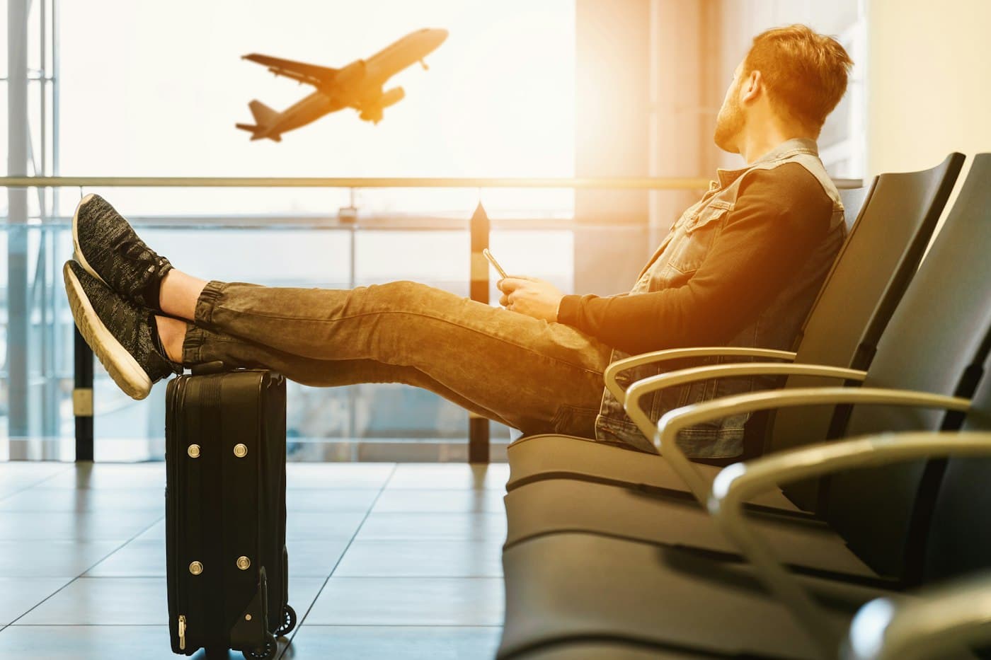 A traveller resting on luggage at an airport gate, watching a plane take off